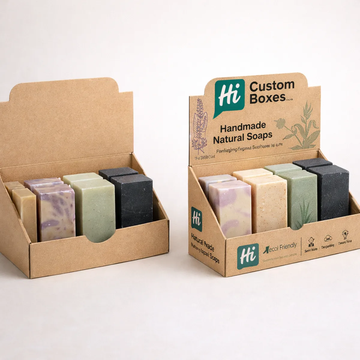 Two cardboard counter-top display units filled with assorted handmade soap bars, featuring green branding and natural botanical illustrations. class=